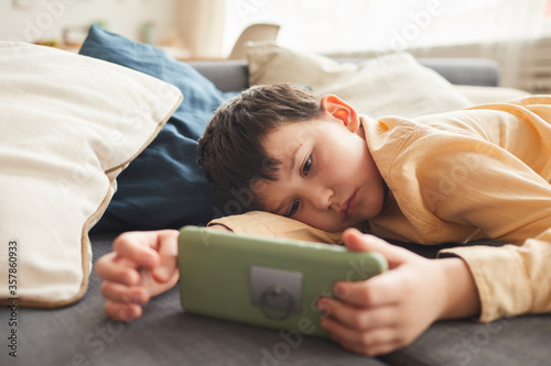 Warm-toned portrait of bored teenage boy using smartphone while lying on bed or couch at home, copy space