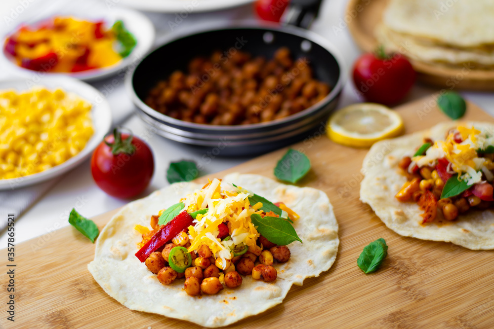 Tortilla tacos with chickpeas, bell peppers, corn, tomatoes, basil leaves, green chilli topped with cheddar and Monterrey Jack. On the background are ingredients, lemons, chickpeas in a small pan