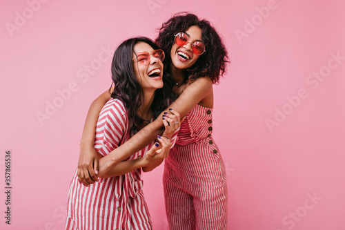Sincere girls with radiant smile pose in striped pink overalls. Summer shot in studio of funny models