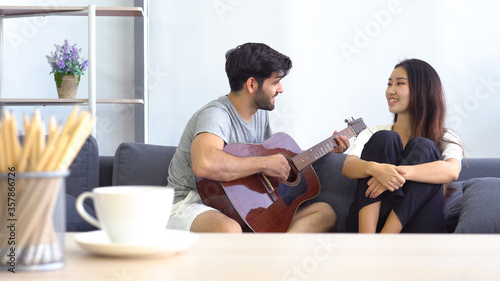 young caucasian man playing guitar and singing song to asian woman in the living room at home. family together and couple relationship concept