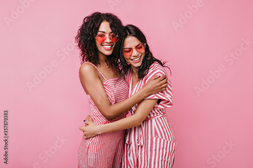 Tanned girls with sincere white smiles rejoice photo shoots. Girlfriends in summer jumpsuits posing on isolated background