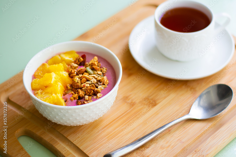 Healthy summer breakfast of a pink smoothie bowl made with frozen banana and raspberry garnished with granola and fresh orange. In the background is black tea Healthy summer breakfast of a pink smoothie bowl made with frozen banana and raspberry garnished with granola and fresh orange. In the background is black tea