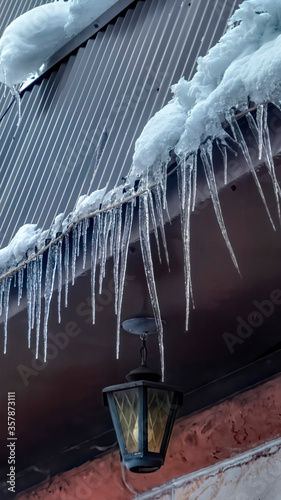 Vertical frame Spiked icicles at the edge of pitched gray roof with clumps of snow in winter © Jason
