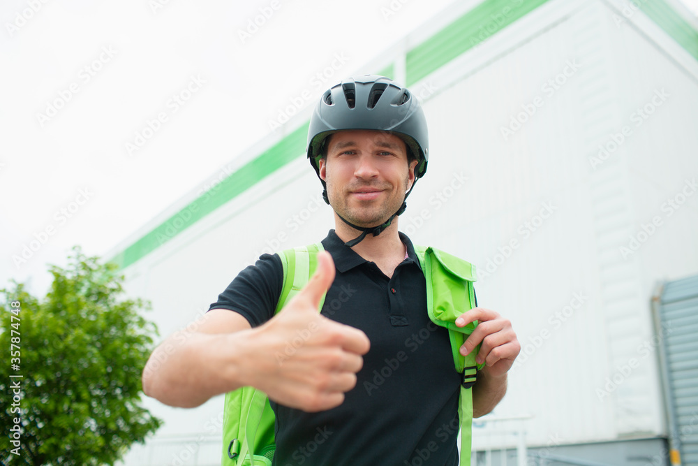 Food delivery concept. The food delivery man uses a smartphone to reach customers faster. Courier has a refrigerator in a green backpack. He is looking at the camera and holds his thumb up. Food delivery concept. The food delivery man uses a smartphone to reach customers faster. Courier has a refrigerator in a green backpack. He is looking at the camera and holds his thumb up.