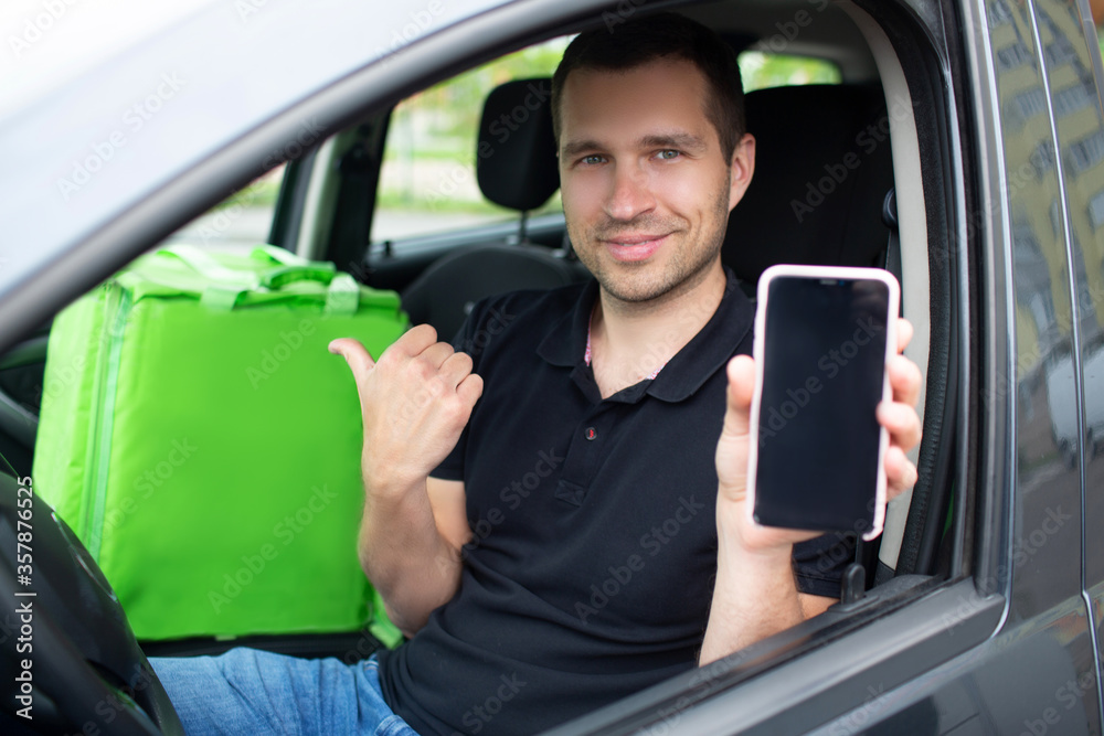 The food delivery man has a green fridge backpack. He sits in a car. He shows the screen of a smartphone with a food delivery application, looks at the camera The food delivery man has a green fridge backpack. He sits in a car. He shows the screen of a smartphone with a food delivery application, looks at the camera