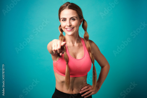 Sexy skinny blonde fitness girl wearing sport clothes with braids smiling and looking at camera while points with her finger on a blue background