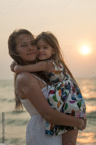 Smiling mother and beautiful daughter having romantic evening on the beach during sunset. Portrait of happy woman hugging to cute little girl on the sea. Portrait of happy blonde kid embracing her mom