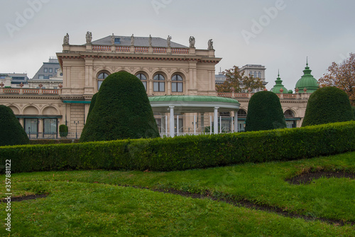 Beautiful city park with green lawn in Vienna, Austria. A building with sculptures on the roof in the background.