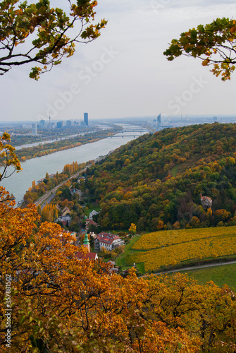 View of the Danube River and the city of Vienna, Austria on an autumn day. Bright yellow and green leaves on trees in the park, skyscrapers and bridges on the horizon. © Hanna