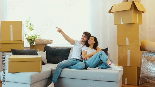Young couple move into new apartment. Beautiful couple sit together on sofa and having fun. Guy pointing forward. Camera moves to the back. Making plans for apatment and furniture. photo