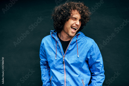 Cheerful young man with curly hair blinking with an eye to the camera. Happy male resting in the street on a rainy day. Happy guy poses against black wall.