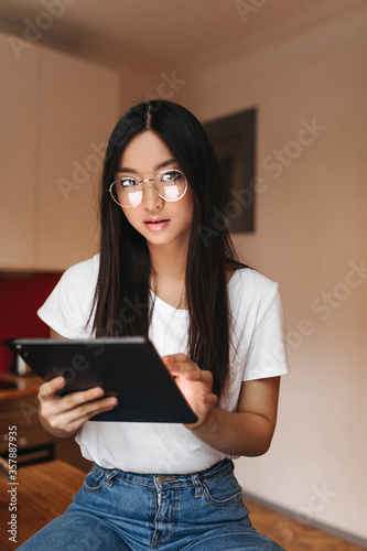 Brunette woman in glasses is posing with tablet. Asian in white top looking at camera