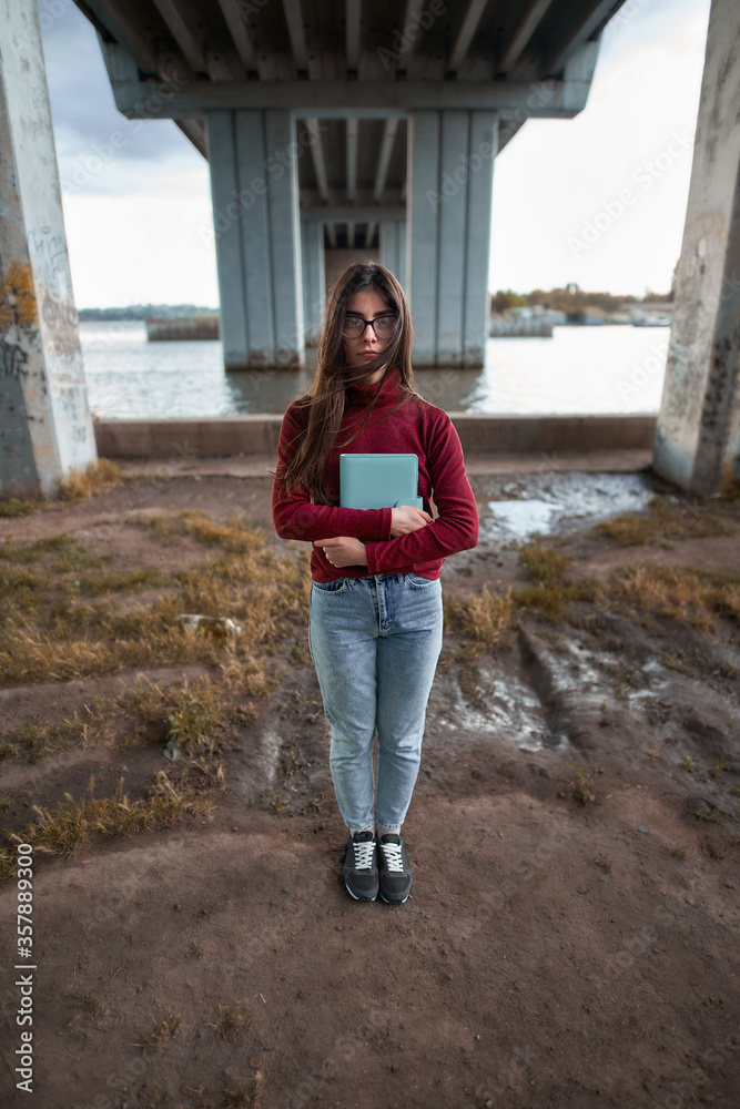 girl in the glasses hold notebook under the bridge urban construction. young female student waiting