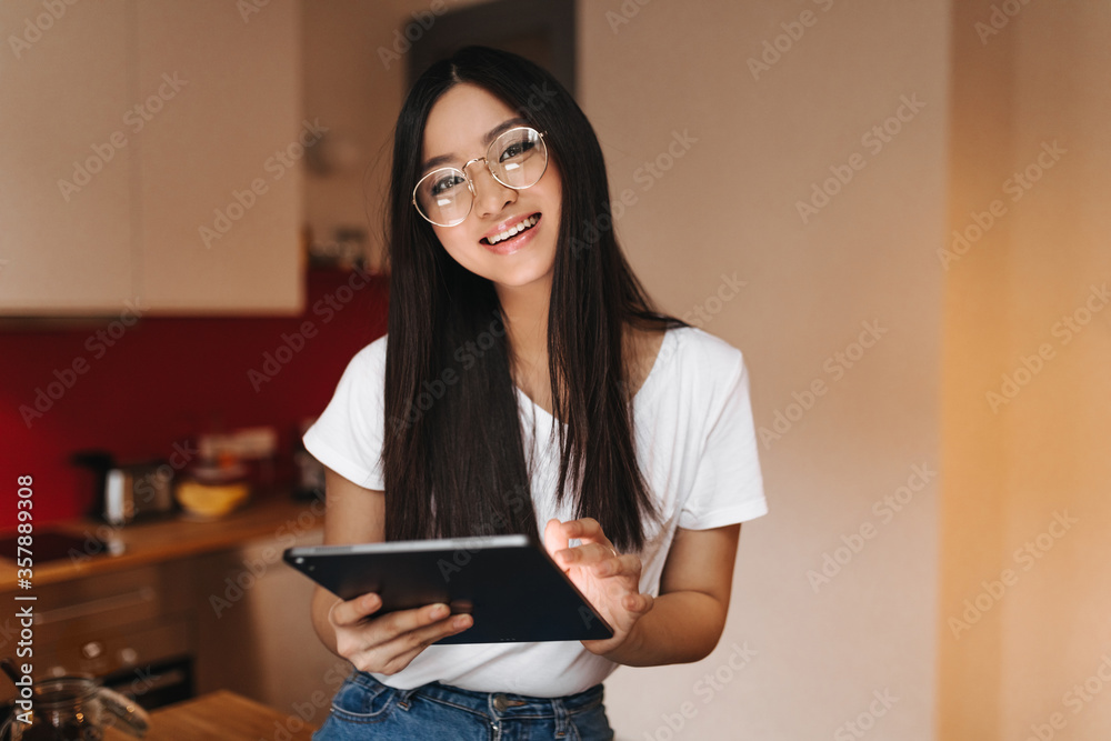 Woman with dark long hair is smiling and holding tablet. Girl in white top and glasses posing in kitchen Woman with dark long hair is smiling and holding tablet. Girl in white top and glasses posing in kitchen