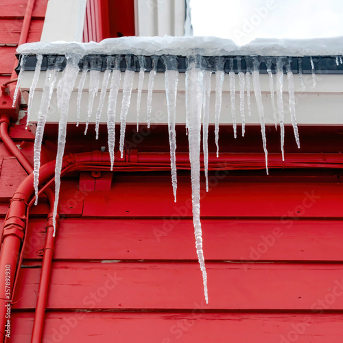 Square Spiked frozen icicles at the roof of home with vibrant red wooden exterior wall © Jason
