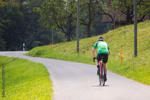 Back shot of unrecognizable cyclist riding bike along road in park alone at a daytime. Stylish male biker in black sport shorts and green T-Shirt cycling on path with trees and house on a background