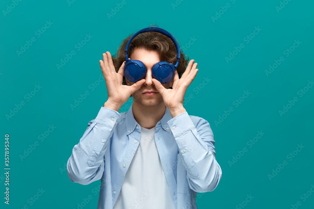 grimacing. Portrait of a young beautiful man wearing white t-shirt and blue shirt cover his eyes by speakers of blue headphones grimacing. Portrait of a young beautiful man wearing white t-shirt and blue shirt cover his eyes by speakers of blue headphones