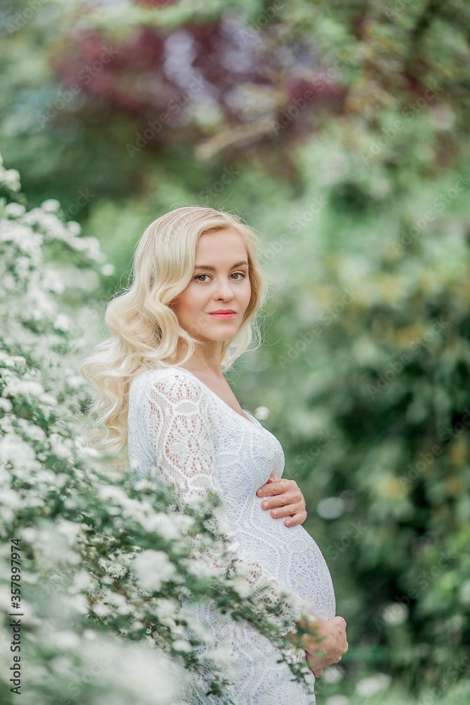 Young beautiful pregnant woman in a lace white dress walks in a flowering park. Portrait of a beautiful pregnant blonde. Spring.