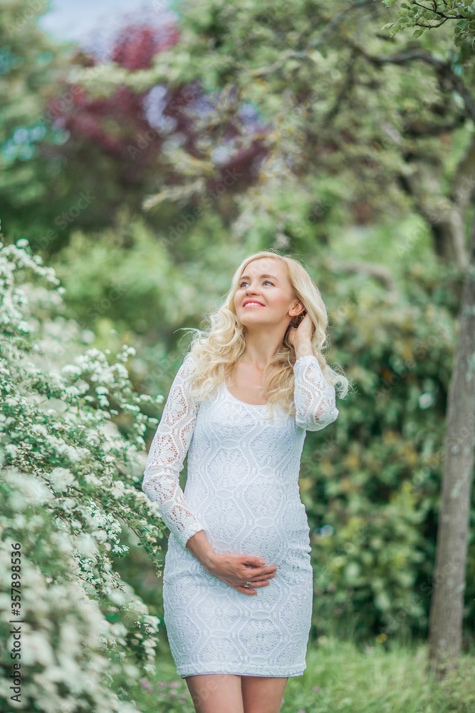 Young beautiful pregnant woman in a lace white dress walks in a flowering park. Portrait of a beautiful pregnant blonde. Spring. Young beautiful pregnant woman in a lace white dress walks in a flowering park. Portrait of a beautiful pregnant blonde. Spring.