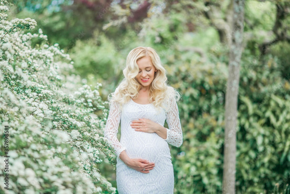 Naklejka premium Young beautiful pregnant woman in a lace white dress walks in a flowering park. Portrait of a beautiful pregnant blonde. Spring.