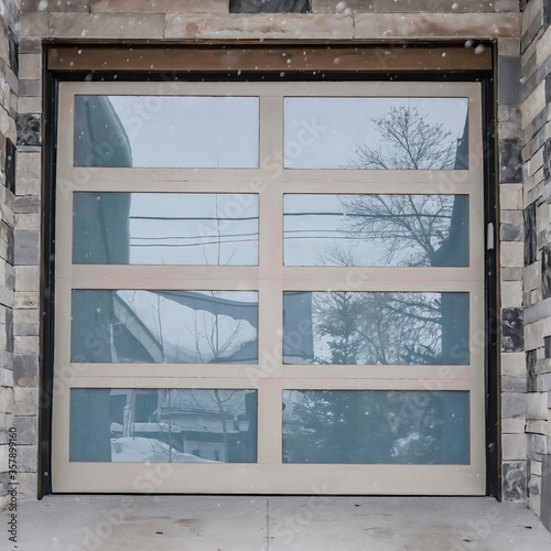 Square frame Snow falling down on home with glass panel garage door and stone exterior wall