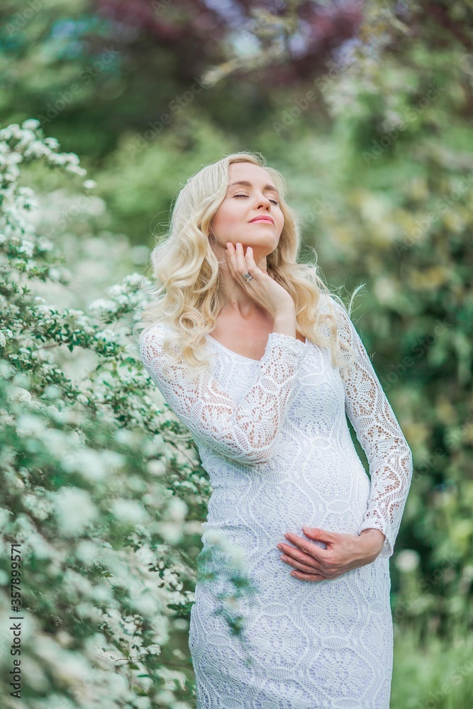 Young beautiful pregnant woman in a lace white dress walks in a flowering park. Portrait of a beautiful pregnant blonde. Spring. Young beautiful pregnant woman in a lace white dress walks in a flowering park. Portrait of a beautiful pregnant blonde. Spring.
