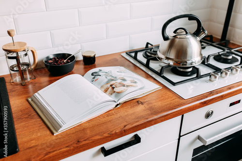 Big cook book on the wooden table. Metallic teapot on the gas oven. White kitchen interior. Wooden complete kitchen with gas oven. photo