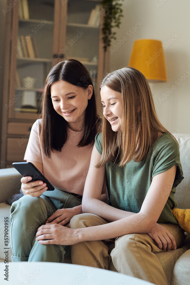 Cheerful mother sitting on sofa and using smartphone while watching funny video with daughter Cheerful mother sitting on sofa and using smartphone while watching funny video with daughter