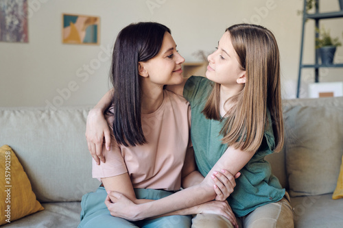 Loving teenage daughter sitting on sofa and embracing mother while looking at her face