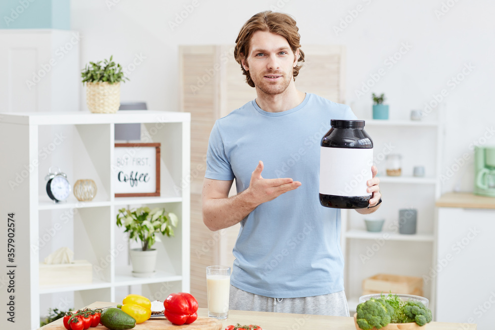 Portrait of sporty young man pointing at bottle with sport nutrition and looking at camera while standing in the kitchen Portrait of sporty young man pointing at bottle with sport nutrition and looking at camera while standing in the kitchen
