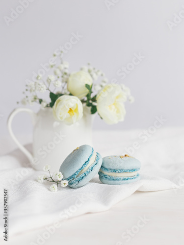 Tasty blue  french macarons and jar with cream roses on a white wooden background.