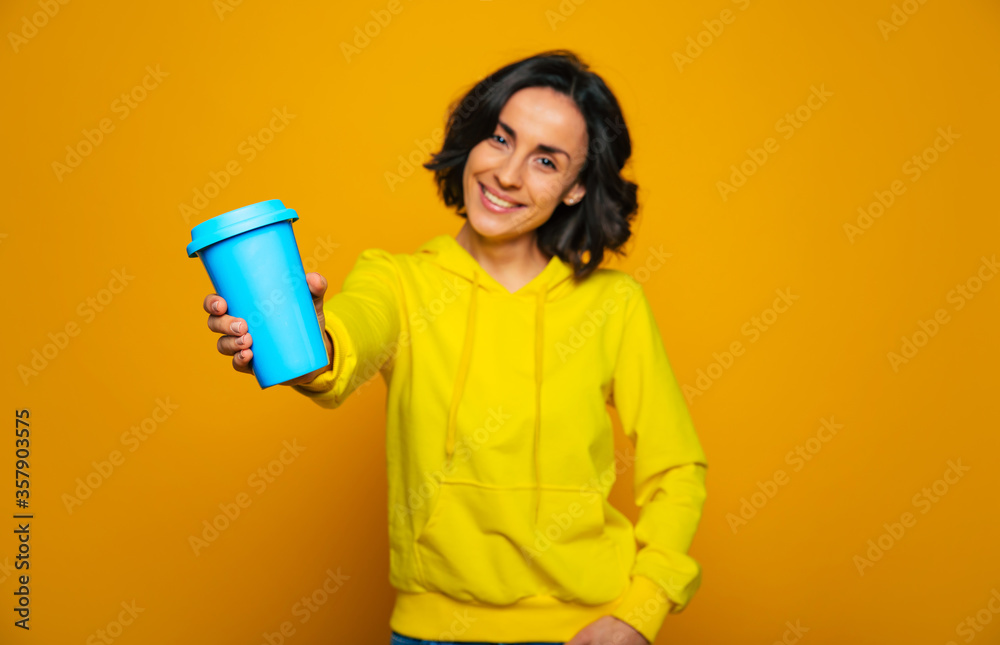 Try my drink. Half-length photo focused on thermal mug, holded on outstretched arm by attractive woman, dressed in a yellow hoodie, offering to try her drink. Try my drink. Half-length photo focused on thermal mug, holded on outstretched arm by attractive woman, dressed in a yellow hoodie, offering to try her drink.