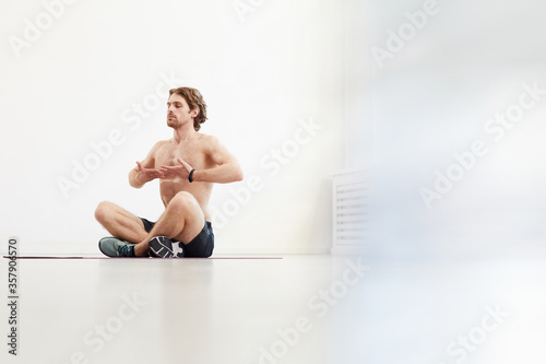 Muscular shirtless man sitting on the floor and doing breathing exercises against the white background