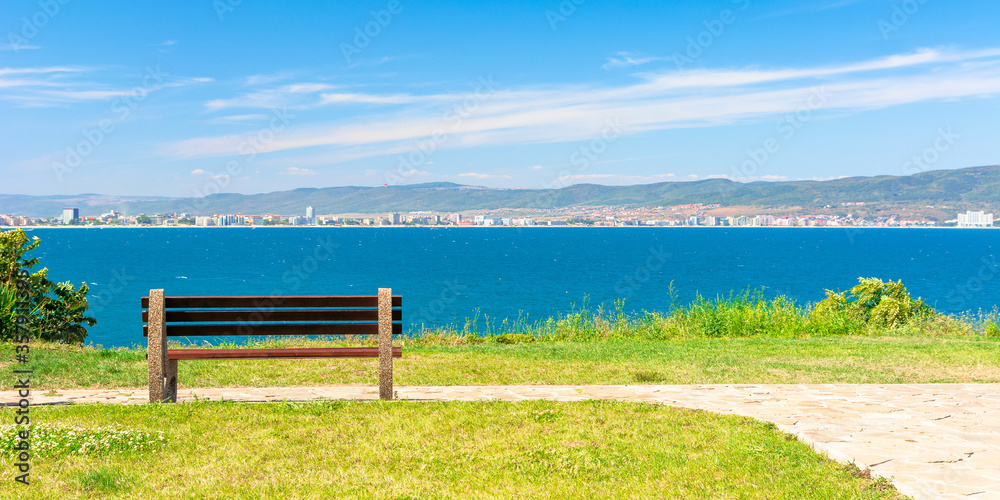 bench on the sunny beach shore. beautiful view from paved footpath on the seaside. city and mountain in the distance beneath a blue sky with clouds bench on the sunny beach shore. beautiful view from paved footpath on the seaside. city and mountain in the distance beneath a blue sky with clouds