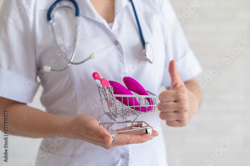 The faceless gynecologist recommends buying a clitoral vaginal vibrator to maintain women's health. The doctor holds a mini trolley and a masturbator for vivid orgasms. Gesture thumb. photo