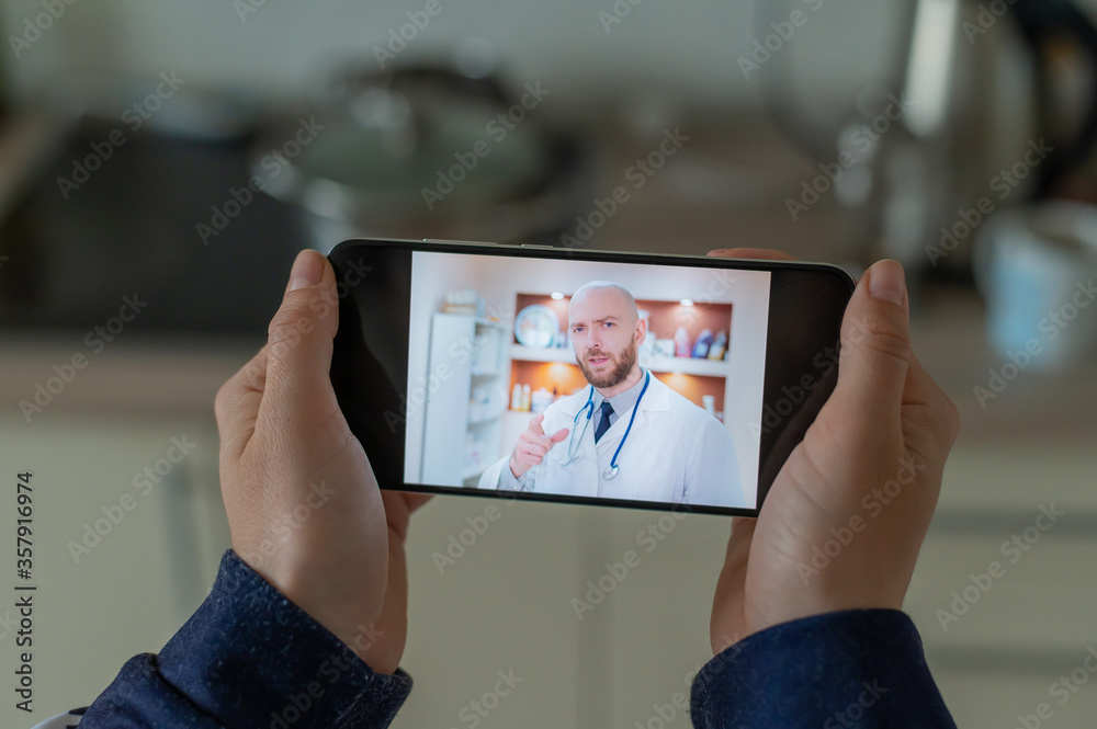 A serious bearded doctor communicates with a patient via a video call on a smartphone. Online medicine concept. Woman in the kitchen takes therapist remote consultation on mobile.