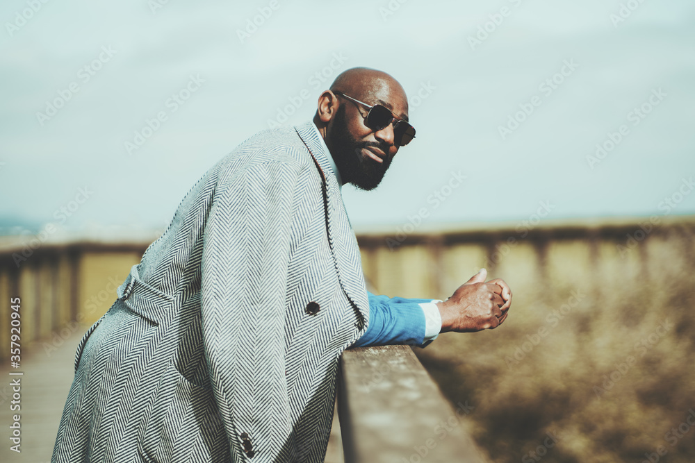 Portrait of a handsome bearded bald African man in sunglasses and coat leaning on a wooden fence outdoors, selective focus, shallow depth of field, a copy space place on the right for an ad message