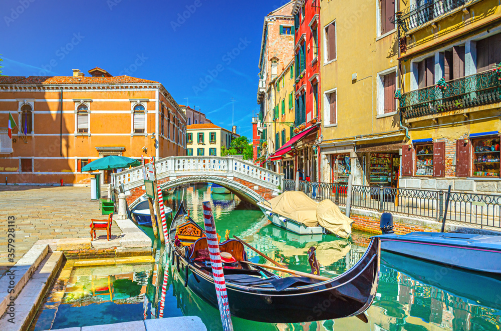 Venice cityscape with gondola and motor boats moored on narrow water canal Rio dei Frari, colorful buildings and stone bridge, Veneto Region, Northern Italy, blue sky background in summer day