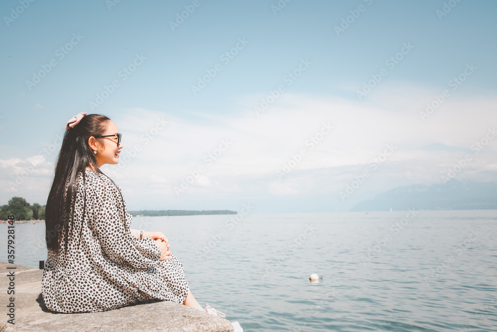 Beautiful Asian woman with white skin and long black hair Wearing black glasses. feeling happy to Sit on stone and watching the lake view On a clear sky day, Tourism and summer vacations. Beautiful Asian woman with white skin and long black hair Wearing black glasses. feeling happy to Sit on stone and watching the lake view On a clear sky day, Tourism and summer vacations.