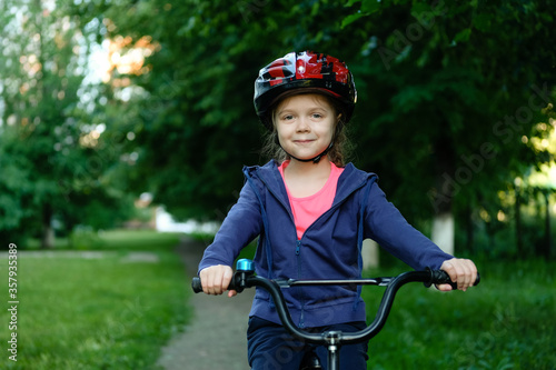 Little girl learns to ride bike in park near home. Portrait of a cute kid on bicycle. Happy smiling baby in helmet on cycling.