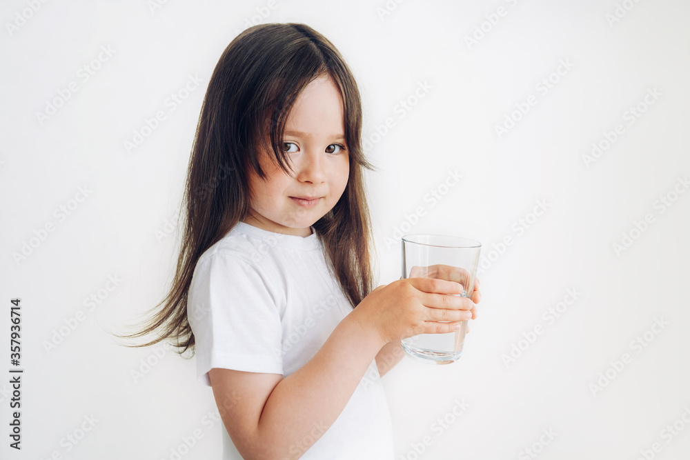 A child in a white T-shirt drinks water. Little girl stands with a glass of water A child in a white T-shirt drinks water. Little girl stands with a glass of water