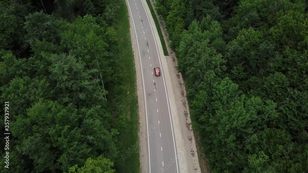 Drone point of view, tracking mode: aerial view flying over two lane countryside forest road with orange car moving green trees of dense woods growing both sides. Car driving along the forest road. Drone point of view, tracking mode: aerial view flying over two lane countryside forest road with orange car moving green trees of dense woods growing both sides. Car driving along the forest road.