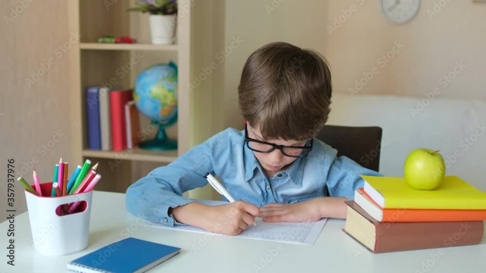 Child Little Boy Kid schoolboy in glasses learning writing homework in kitchen room home, studying making notes sitting at table. Elementary education, children, people, school concept, 4 K slow-mo