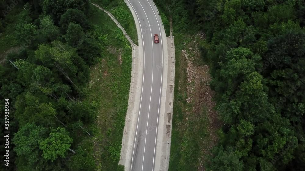Drone point of view, tracking mode: aerial view flying over two lane countryside forest road with orange car moving green trees of dense woods growing both sides. Car driving along the forest road.