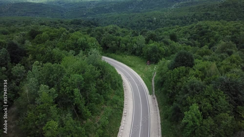 Drone point of view, tracking mode: aerial view flying over two lane countryside forest road with orange car moving green trees of dense woods growing both sides. Car driving along the forest road.