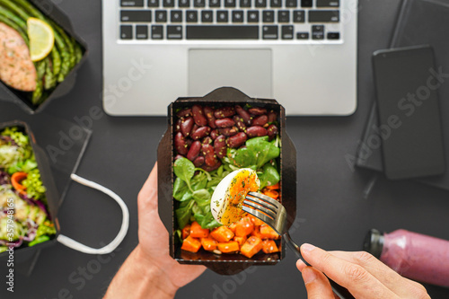 Male hands hold egg on fork vegan food delivery box having lunch at work from home office. Business man employee eats take away daily healthy vegetarian meal at workplace over table flat lay top view.