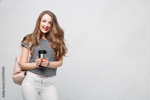 Pretty and stylish brunette girl with red lips, long wavy hair after beauty salon and shopping, posing at studio, holding thermos with warm coffee or tea. Sensuality fashionable woman looking down.