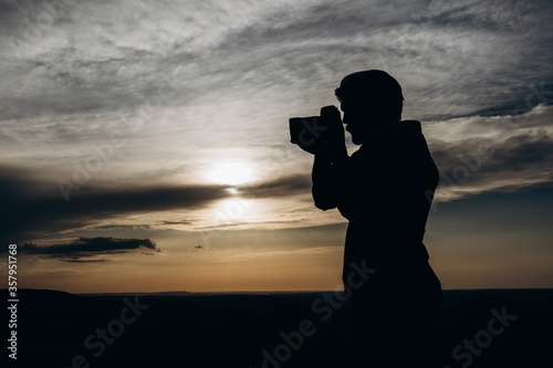 Bearded photographer in silhouette using professional digital camera for making photos of sunset. Mature man spending free time for favorite hobby on fresh air.
