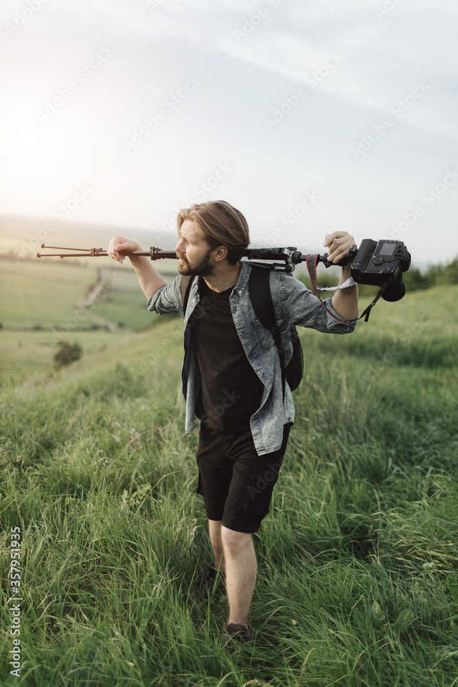 Fototapeta premium Tired bearded man in summer outfit holding tripod with digital camera on shoulder while climbing on high hill. Concept of professional photography outdoors