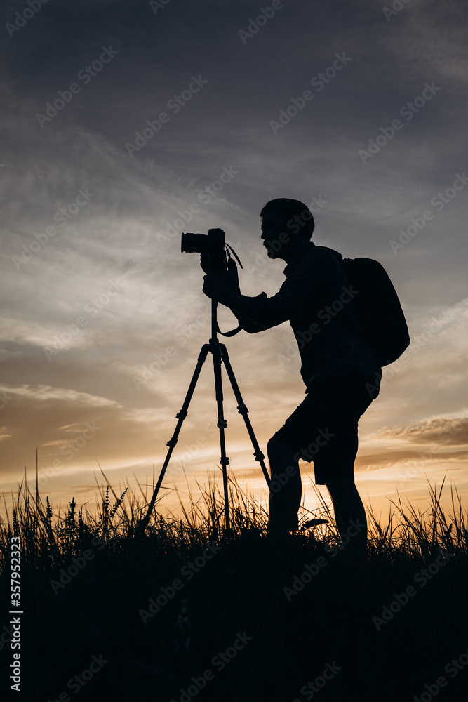 Experienced nature photographer in silhouette using tripod and digital camera for shooting amazing sunset. Concept of working process and environment.